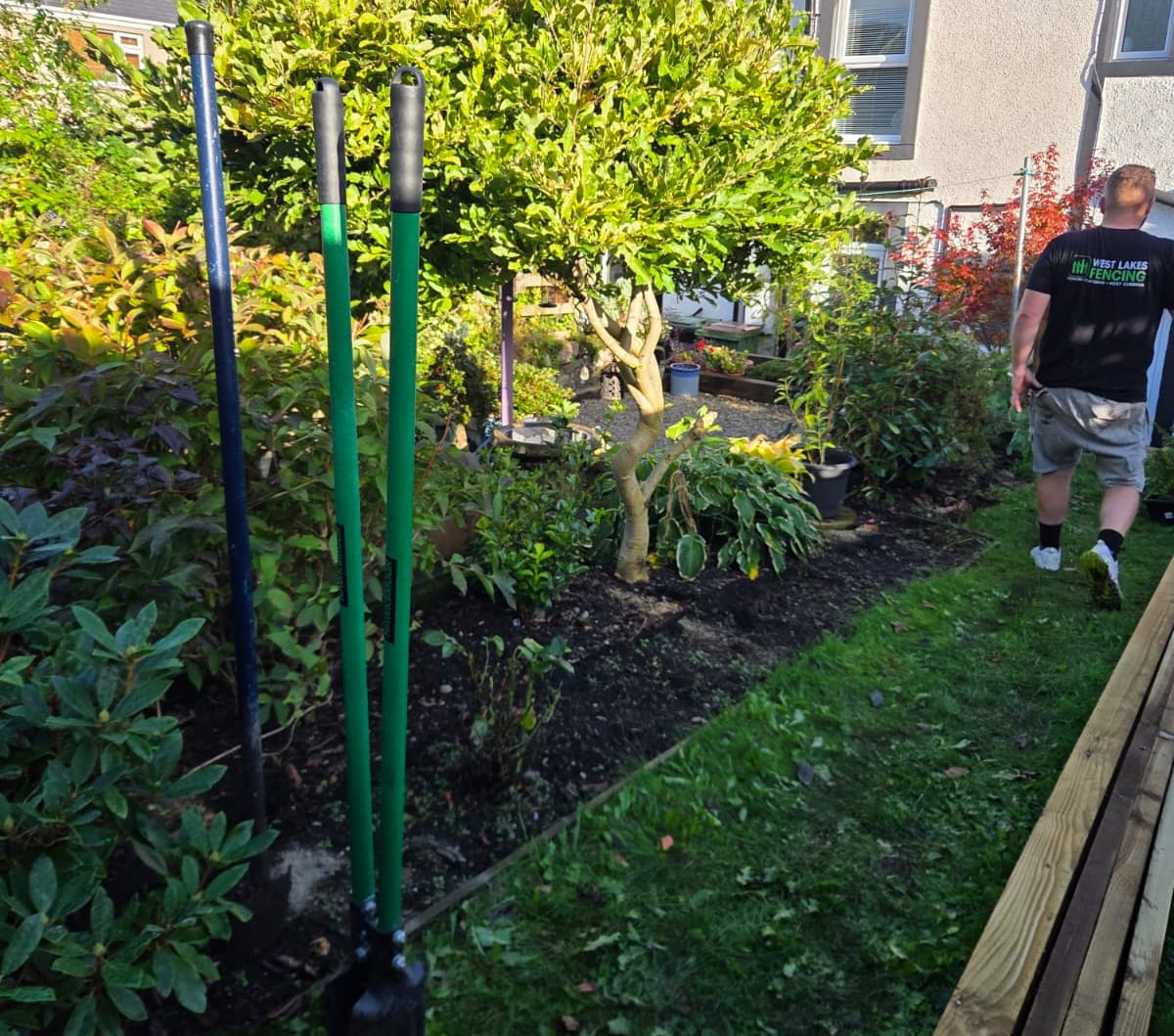 Post hole digger and digging bar standing in a garden border with timber stacked on the lawn